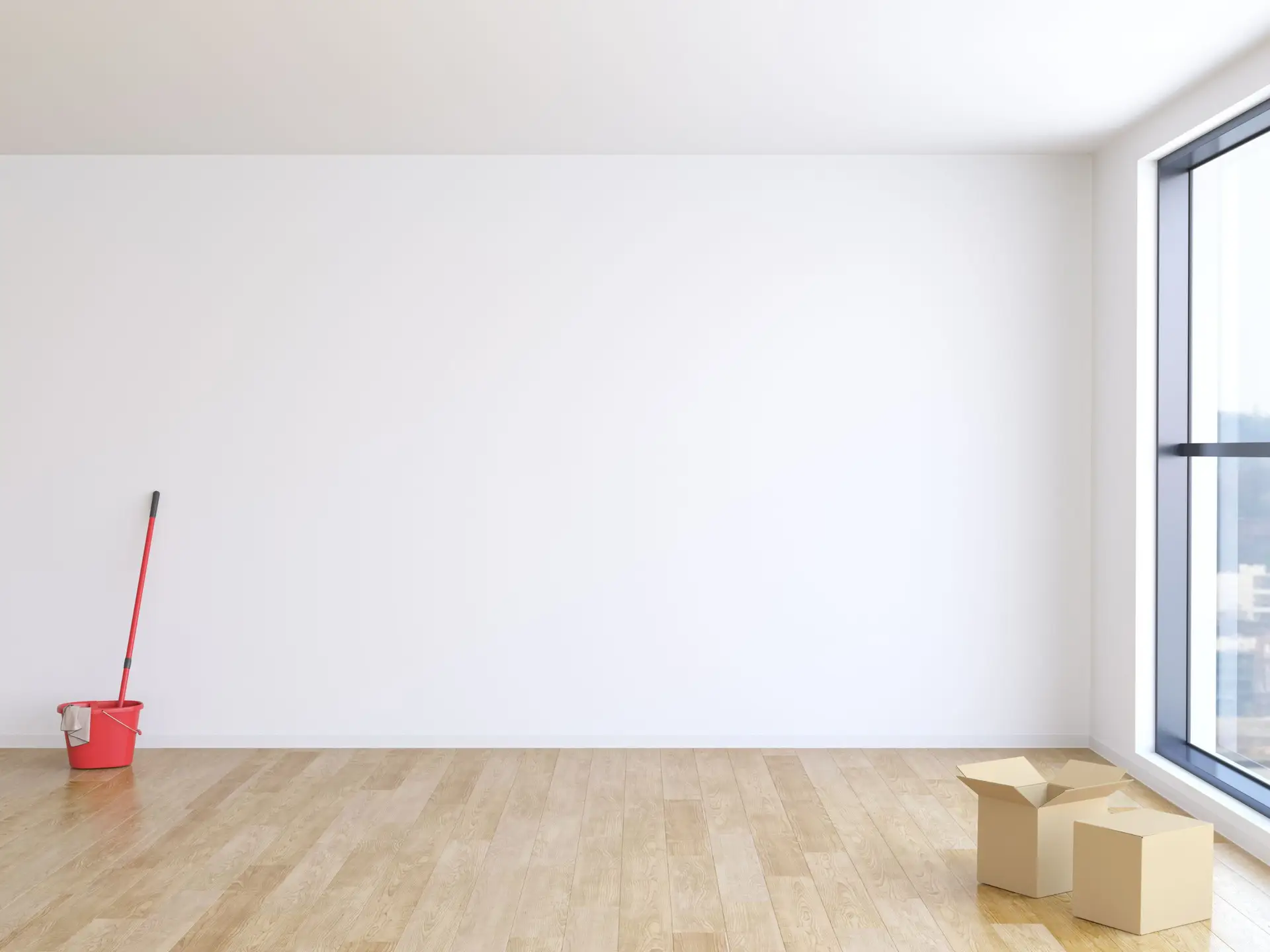 An empty room with wooden floors, a large window, a red mop and bucket in the corner, and two open cardboard boxes on the floor suggests someone is about to move out.