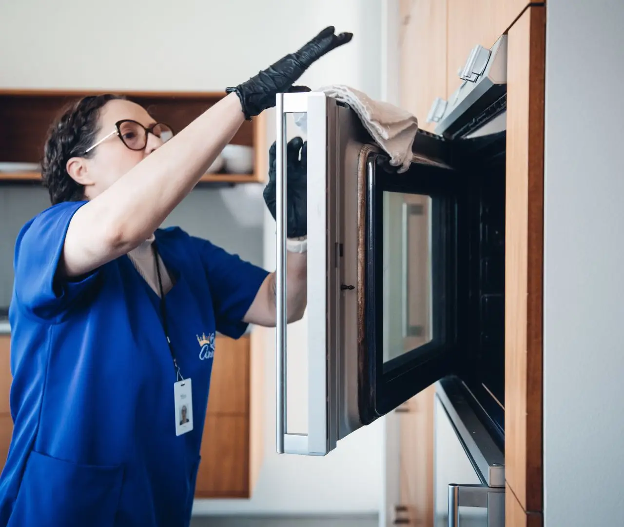 A person wearing gloves and a blue uniform can be seen engaged in recurring cleaning, meticulously scrubbing the inside of an open microwave oven in a kitchen setting.