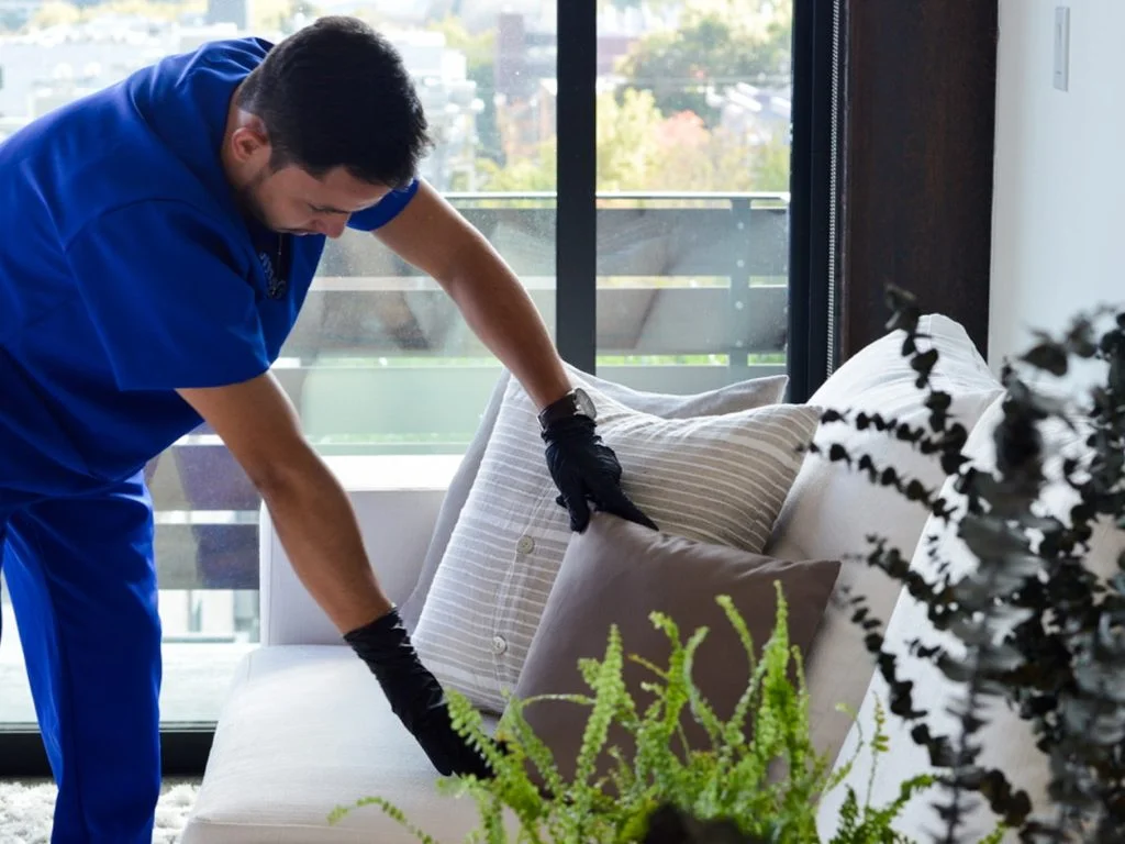 A male worker in blue uniform fluffing pillows on a white couch in a modern living room with urban views, providing house cleaning services.