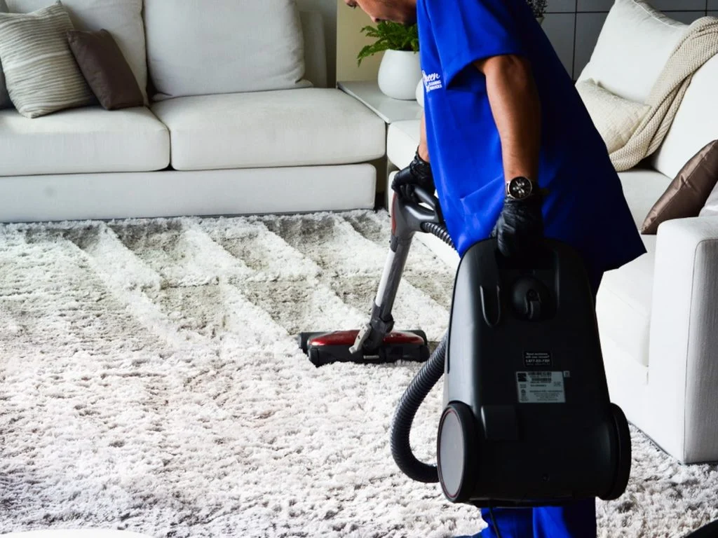A worker in a blue uniform using a portable vacuum cleaner for deep cleaning a white shag carpet in a modern living room.