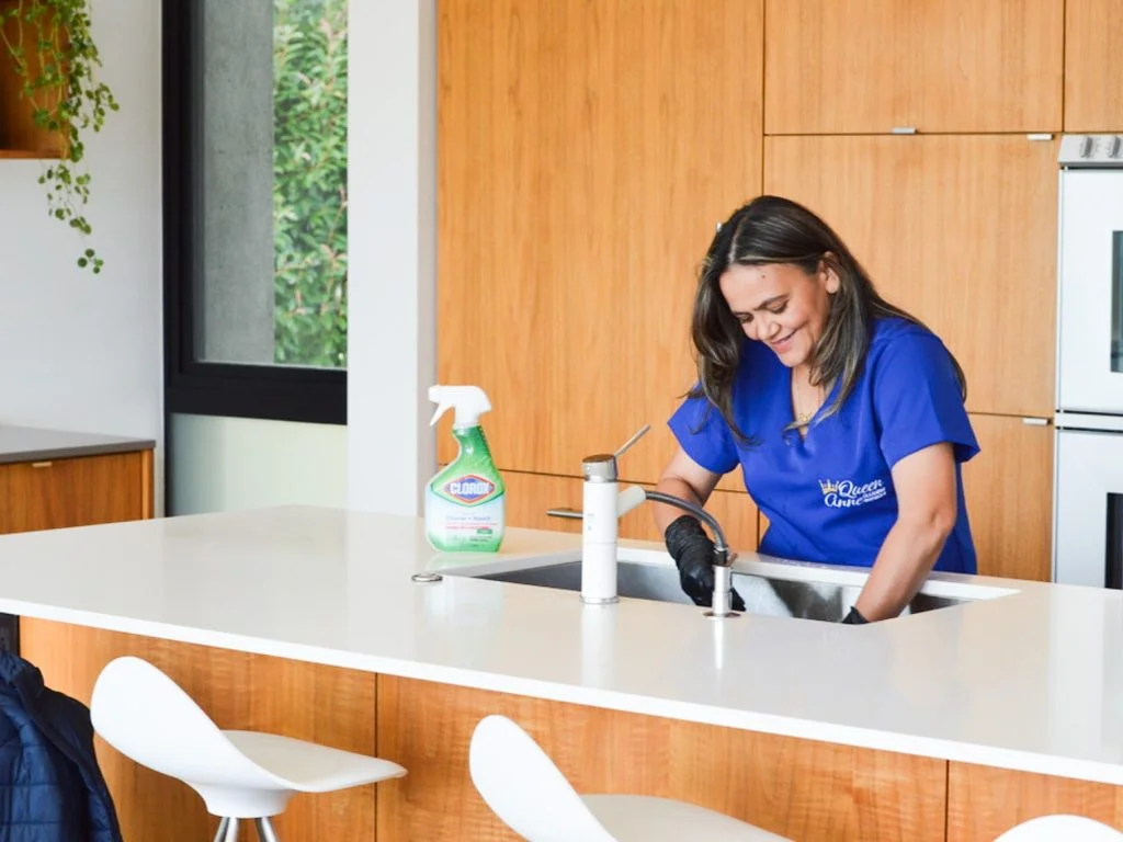 A woman in a blue cleaning uniform smiles while scrubbing a kitchen counter next to a bottle of cleaning spray, offering house cleaning in Seattle.