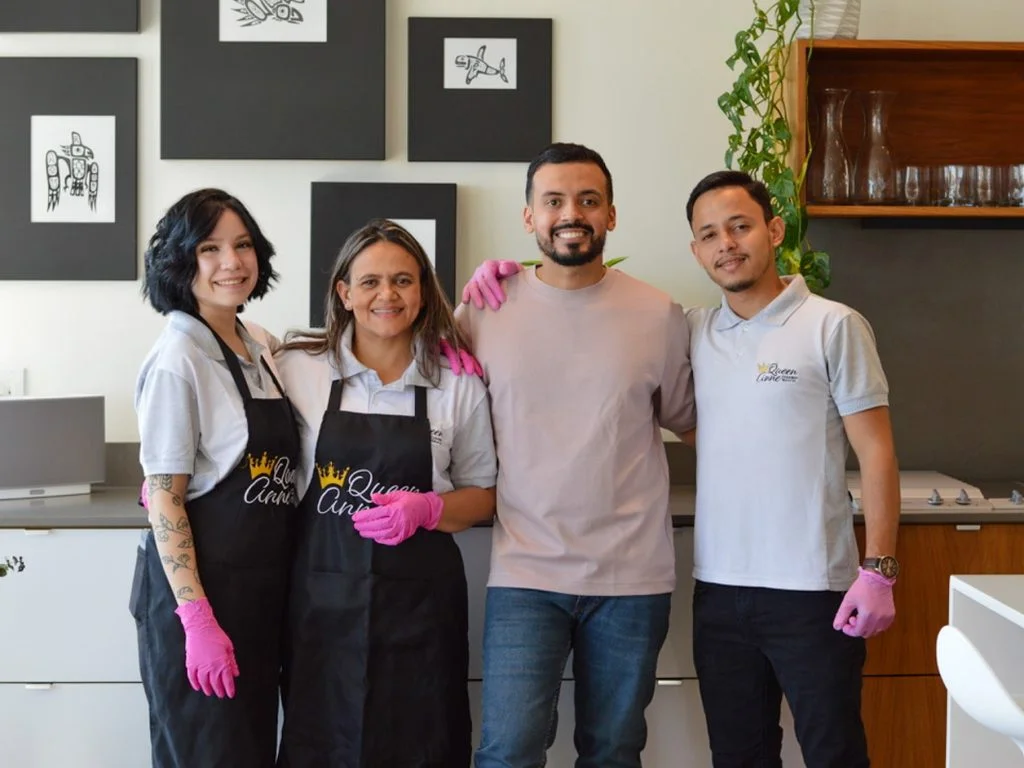 Four people stand together smiling in a kitchen. Two of them wear aprons and pink gloves, likely part of a maid service. Framed artwork is displayed on the wall behind them.