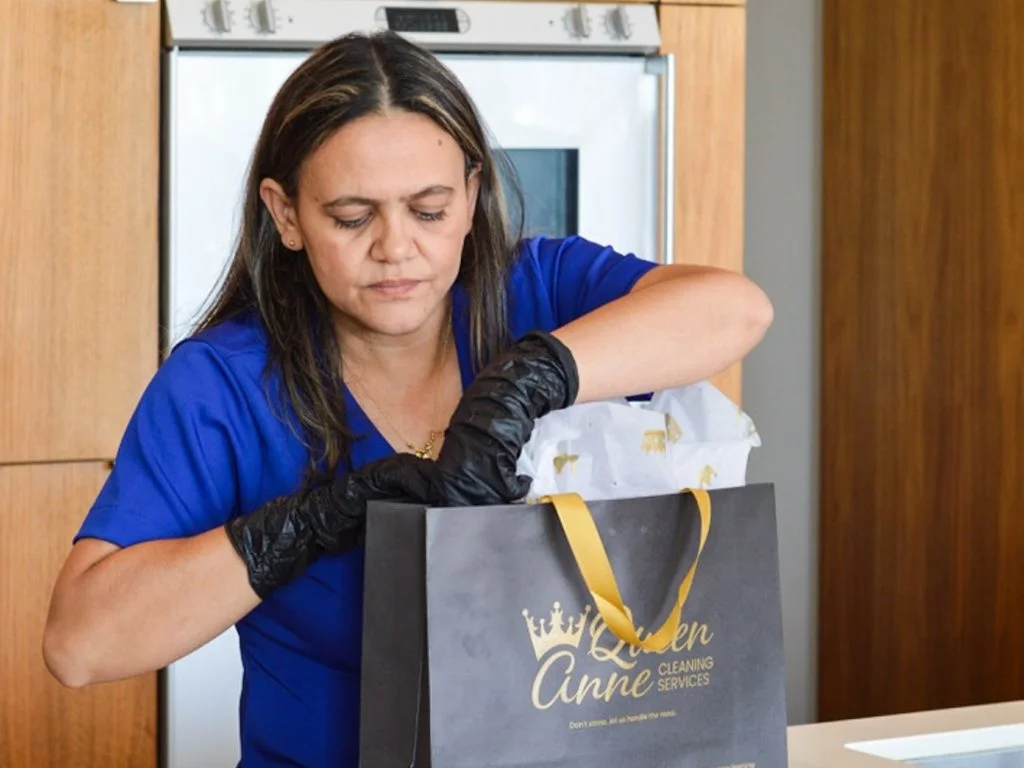 A woman in a blue uniform and black gloves inspects a black gift bag with yellow handles labeled "Queen Anne Cleaning Services" in a kitchen with wooden cabinets, highlighting the premium quality one can expect from house cleaning services near me.