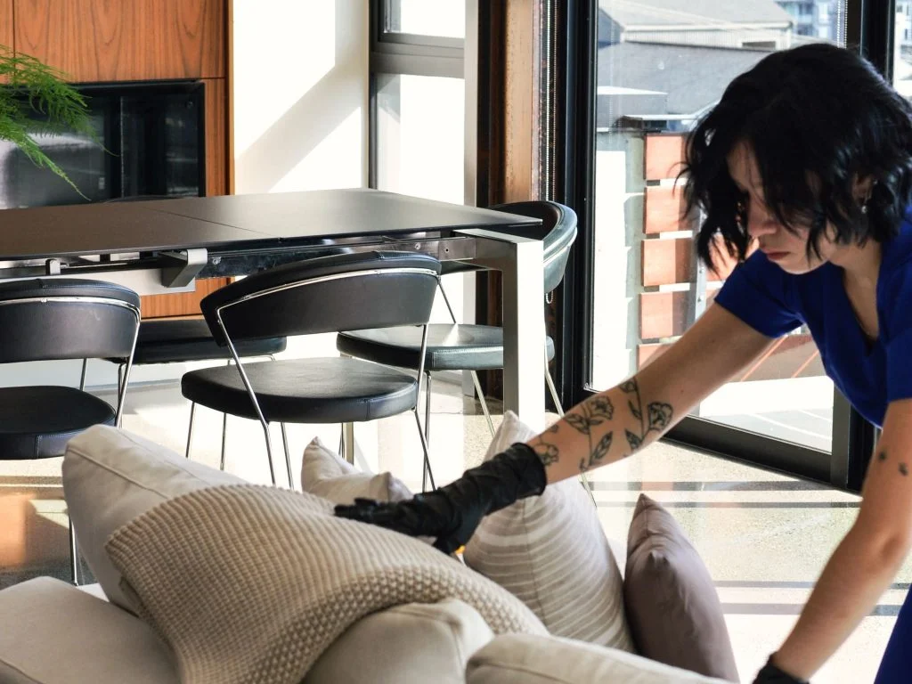 A person with tattoos and black gloves adjusts pillows on a light-colored couch in a modern living room with a large window and black chairs, embodying the efficiency you'd expect from house cleaning services near me.