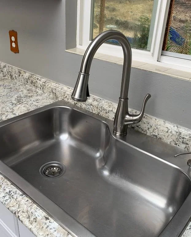 A stainless steel kitchen sink with a gooseneck faucet mounted on a granite countertop, next to a window awaits the sparkle of house cleaning services near me.