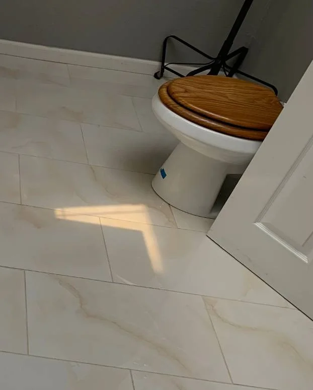 A bathroom floor with beige tiles, a white toilet with a wooden seat, and sunlight shining through a partially open door—perfectly ready for house cleaning services near me to keep it spotless.