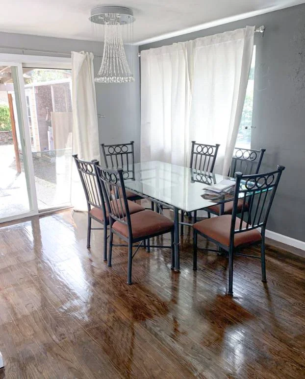 A dining room with a glass table, six metal chairs with brown cushions, a wooden floor, large windows with white curtains, and a hanging chandelier—perfectly maintained by the best house cleaning services near me.