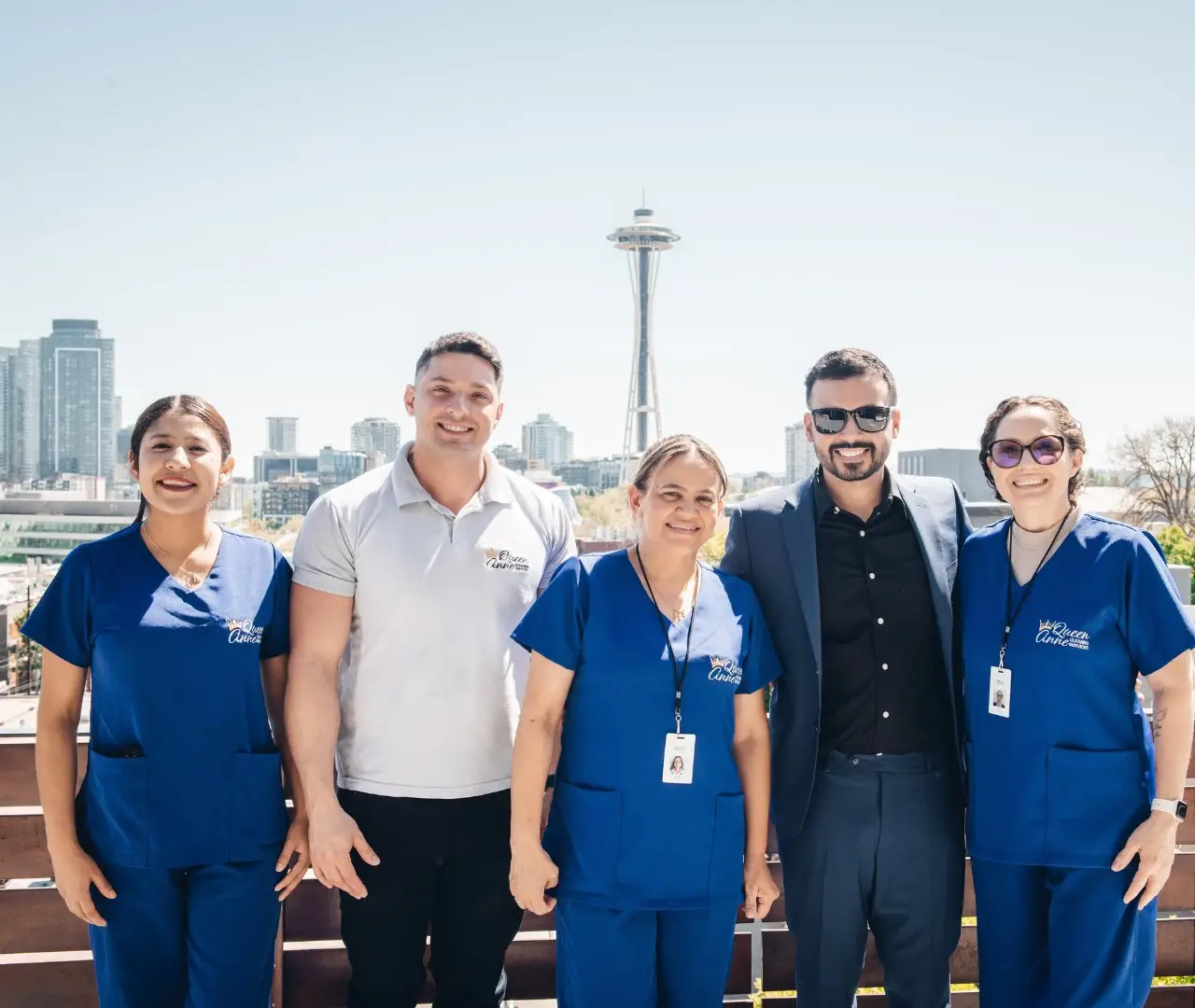 Group of five individuals, three in blue medical scrubs and two in casual/business attire, standing outside with a cityscape and the Space Needle in the background, discussing the best house cleaning services near them.