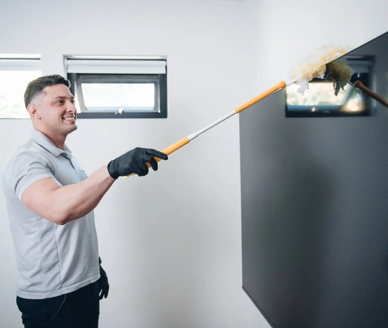 A person wearing a polo shirt and gloves uses a duster to clean a blackboard in a brightly lit room with two windows, showcasing the meticulous attention provided by professional house cleaning services near me.