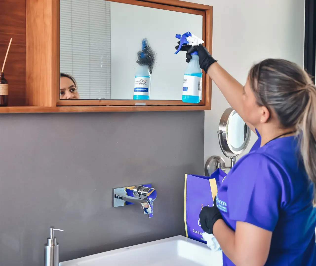 A person wearing a purple uniform and black gloves is cleaning a bathroom mirror with a spray bottle and cloth, demonstrating the high standards of their house cleaning service.