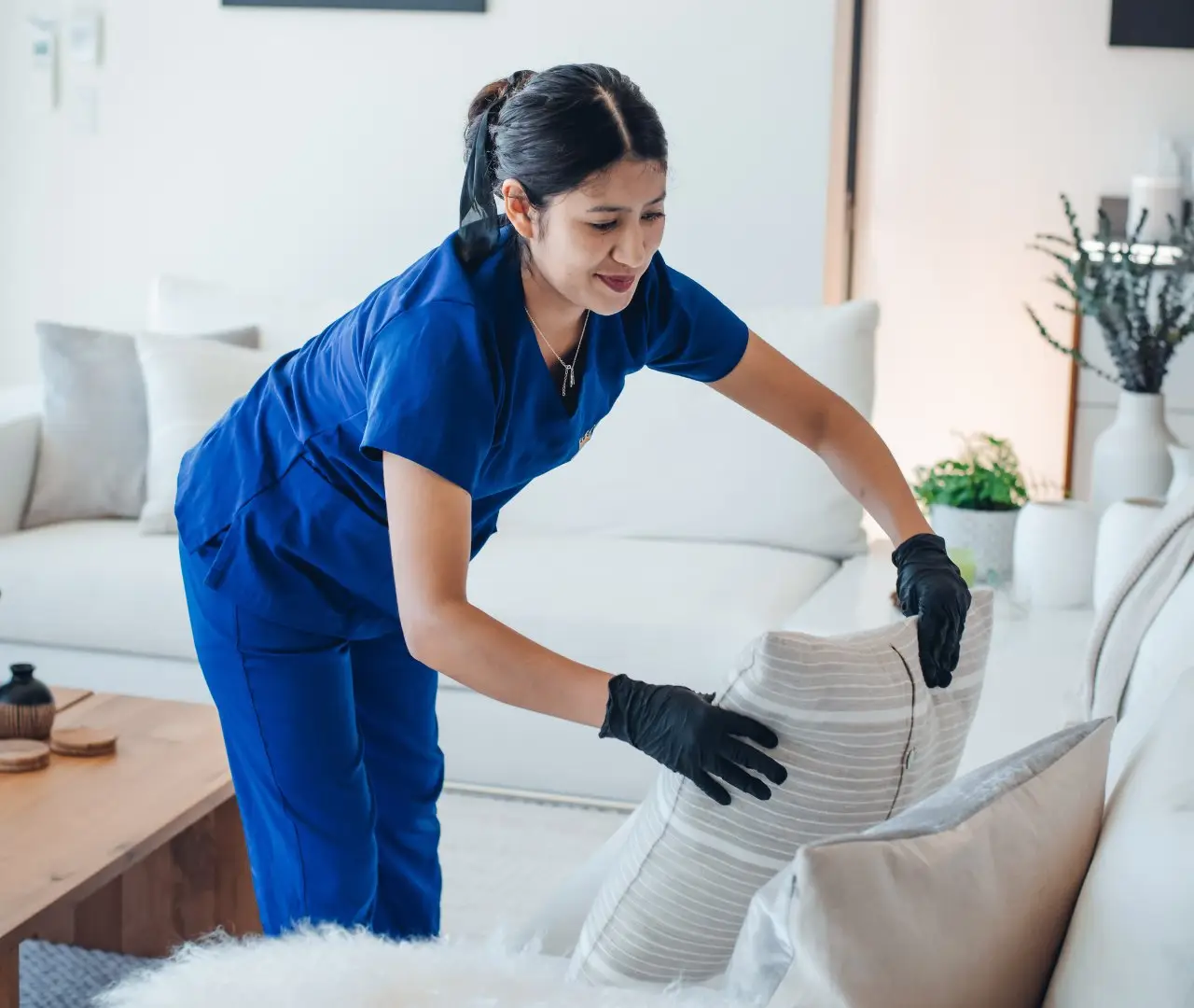 A person wearing blue scrubs and black gloves arranges pillows on a white couch in a well-lit living room, showcasing professional house cleaning services near me.