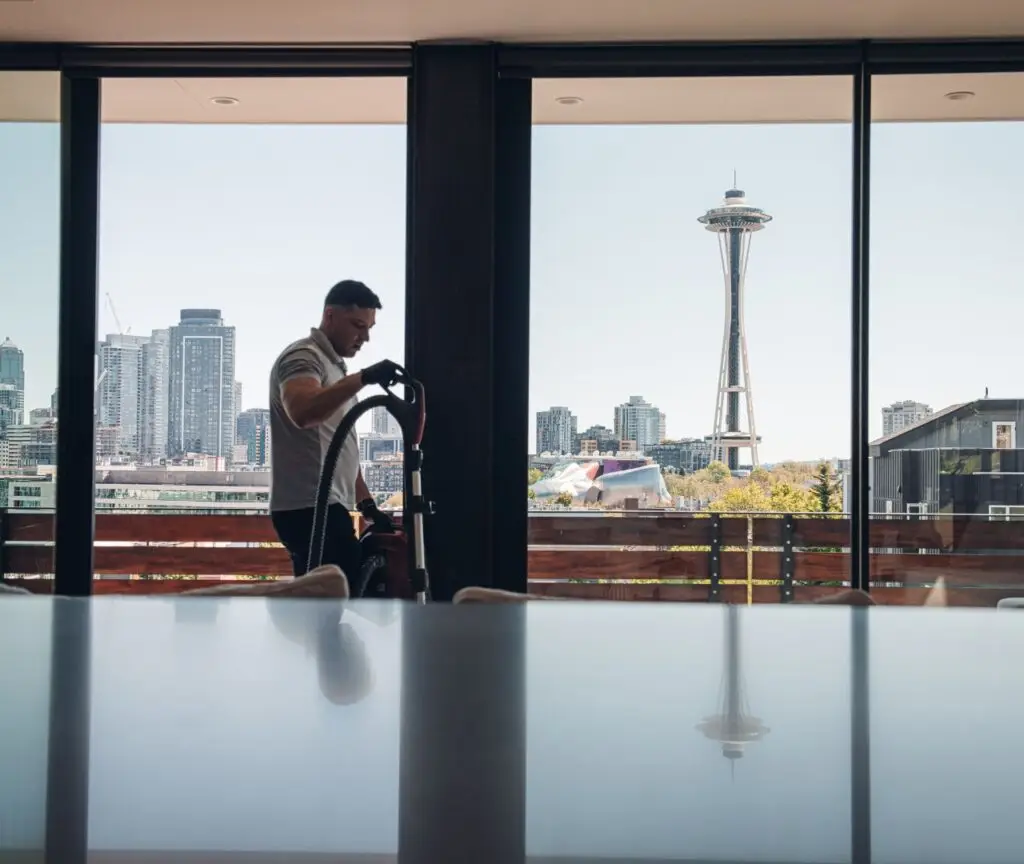 A person vacuums a room with large windows overlooking the Seattle skyline, including the Space Needle, on a clear day—courtesy of Professional Office Cleaners ensuring a pristine workspace.