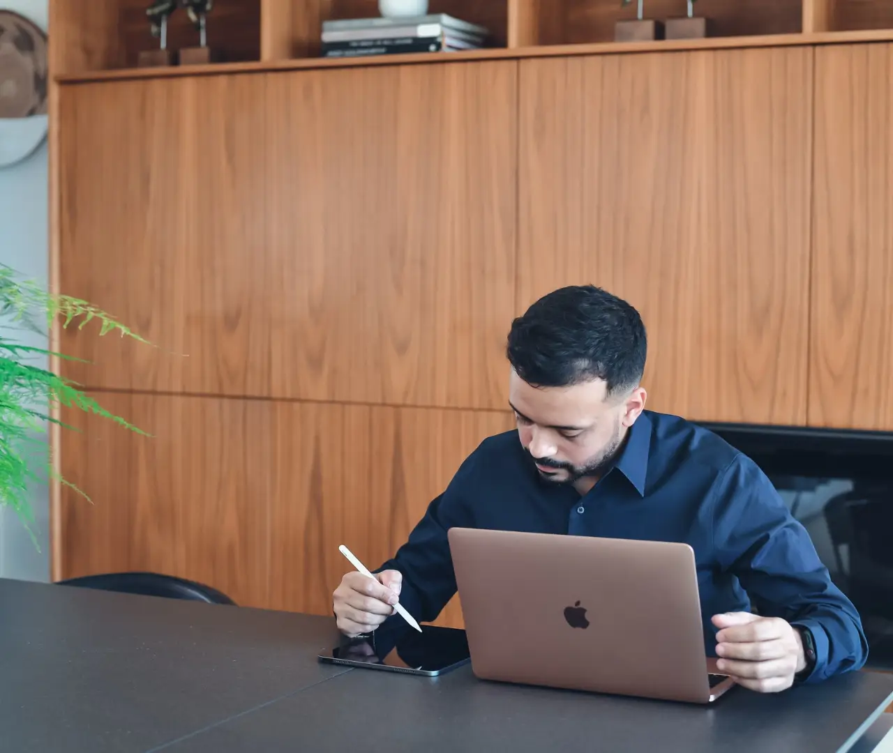A man uses a stylus on a tablet while sitting at a table with an open laptop in front of a wooden cabinet, perhaps searching for nearby maid services to conquer his cluttered home.