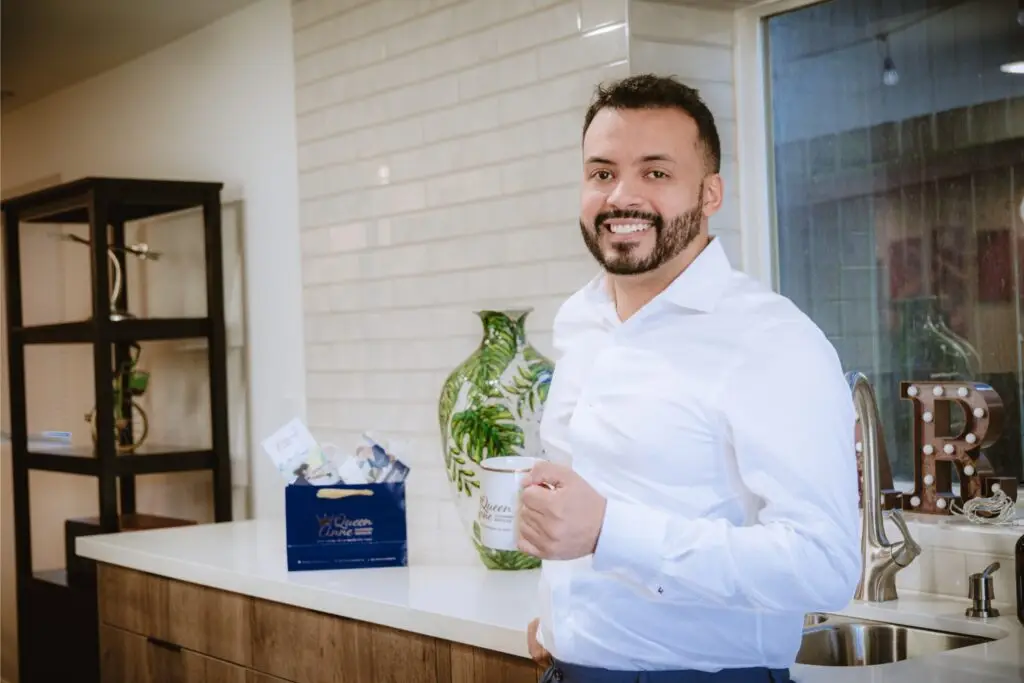 A man in a white shirt stands in a kitchen, smiling and holding a coffee mug. There is a green vase and a blue gift bag on the counter behind him.