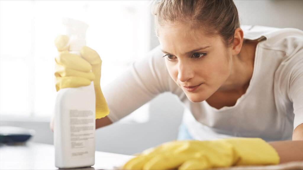 Woman wearing yellow gloves intently cleans a surface with a spray bottle in a well-lit Seattle room, determined to reduce home allergens this spring.