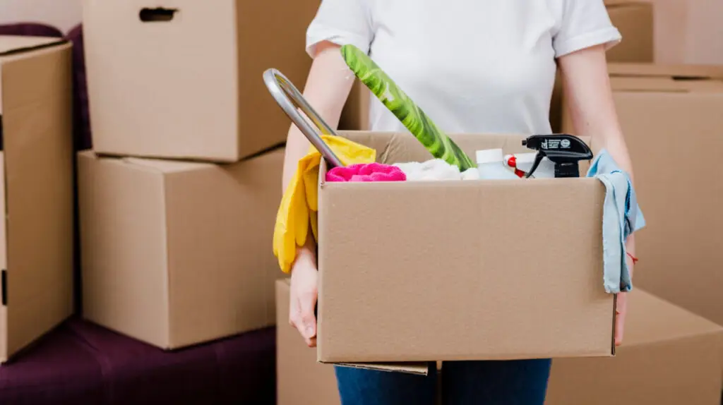 Person holding a box of household items with packed moving boxes in the background, ready to settle in and use helpful move-in cleaning tips for a fresh start.