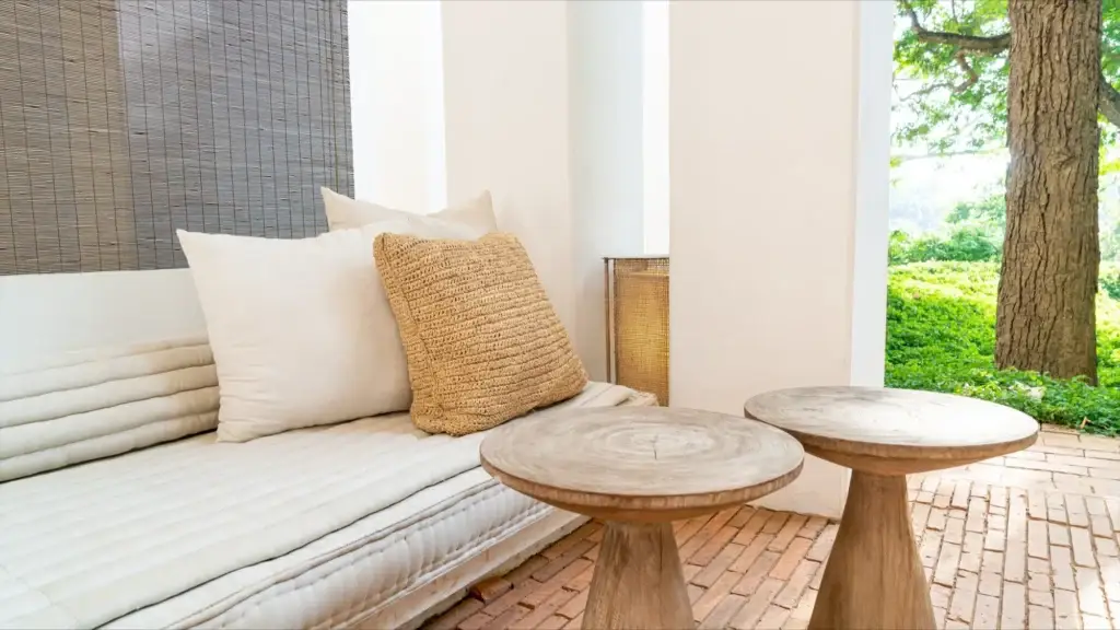 Outdoor patio featuring cozy patio furniture—a cushioned bench with pillows, two wooden tables on a brick floor, and lush greenery outside.
