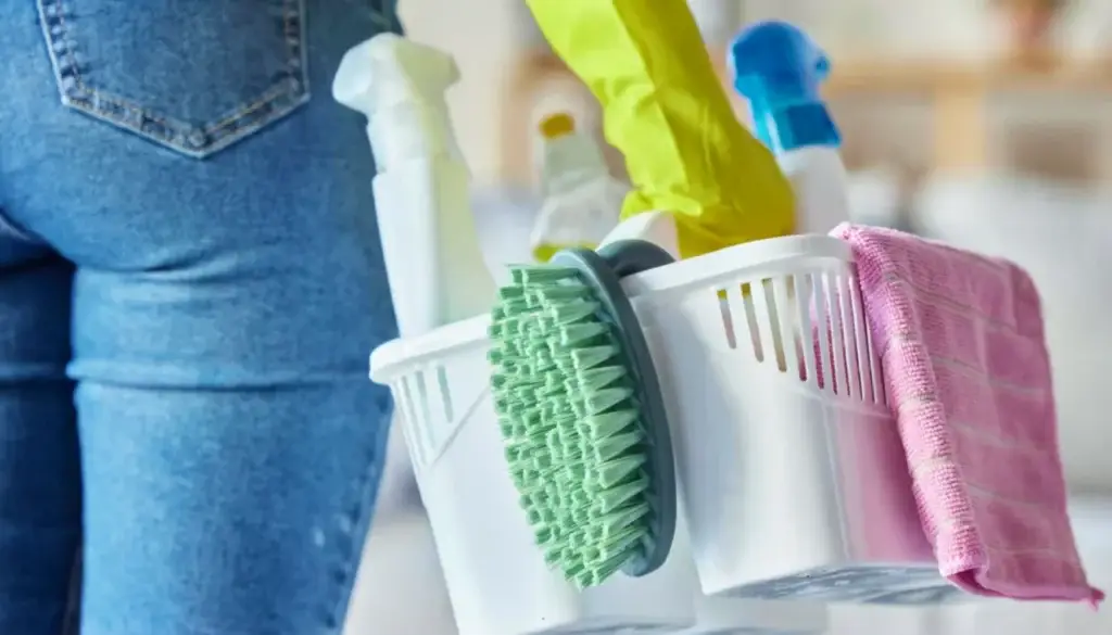 Person holding a cleaning caddy with supplies, wearing yellow gloves and jeans, ready to tackle spring cleaning mistakes.