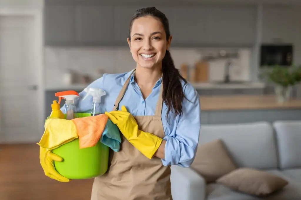 Smiling woman in apron and yellow gloves holds a bucket of cleaning supplies in a living room, ready to deliver professional house cleaning.
