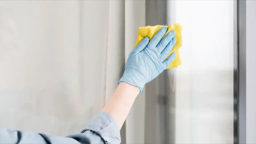 A gloved hand demonstrates how to clean windows by wiping a glass surface with a yellow cloth.