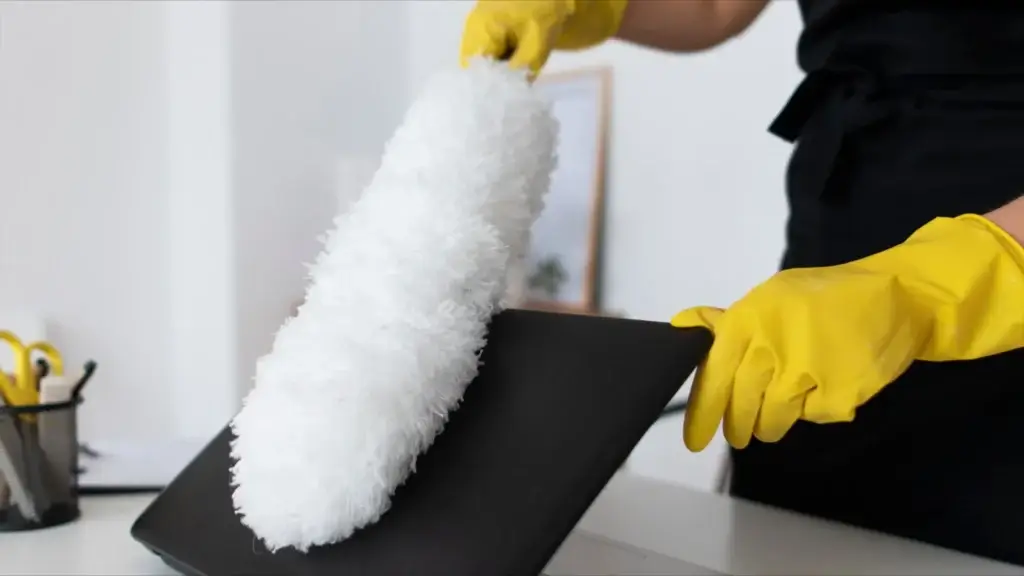 Person wearing yellow gloves dusting a laptop screen with a white duster in a home office cleaning setting.