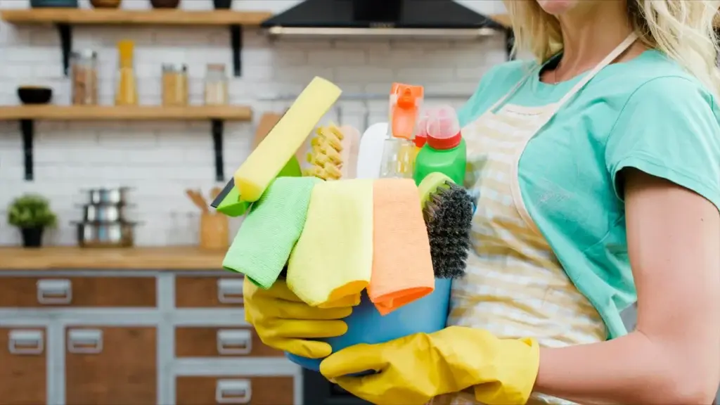 Woman in rubber gloves holding a bucket of zero waste cleaning supplies in a modern kitchen.
