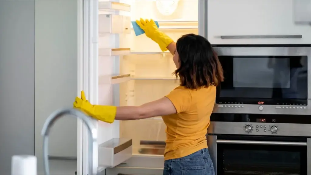 Woman wearing yellow gloves demonstrates how to deep clean a refrigerator, thoroughly cleaning the inside of an open fridge in a modern kitchen.