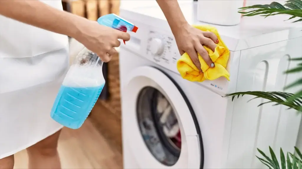A person tackles laundry room cleaning by wiping down a washing machine with a yellow cloth and spray bottle.