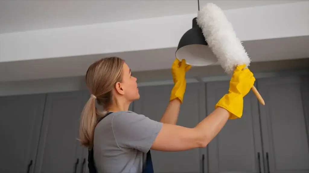Woman wearing yellow gloves dusting a hanging lamp with a white duster in a modern kitchen, showcasing professional Seattle apartment cleaning.