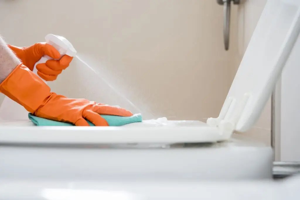Person wearing orange gloves cleaning a toilet lid with spray cleaner and a cloth, ensuring a spotless finish with the best toilet bowl cleaner.