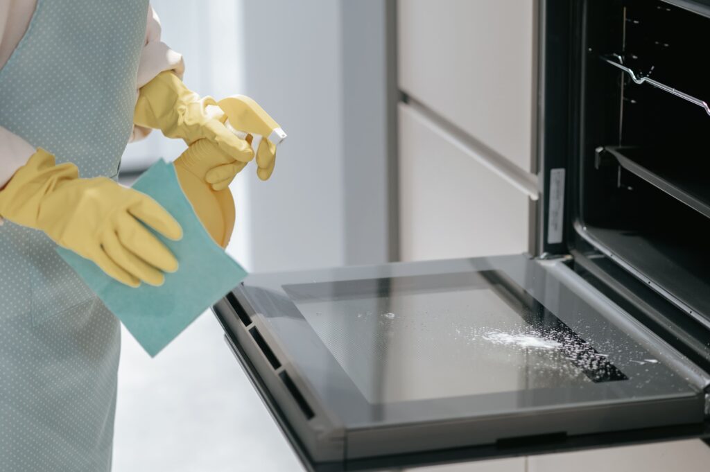 Person wearing yellow gloves cleaning an oven door with a spray bottle and a blue cloth.