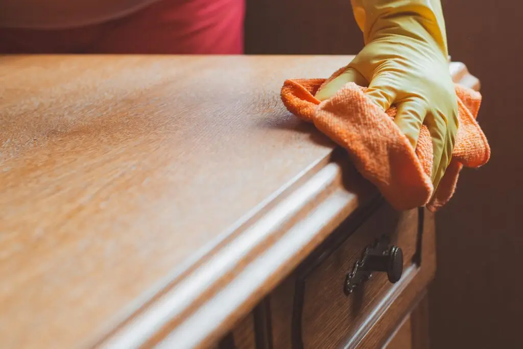 A person wearing a yellow glove wipes a wooden surface with an orange cloth, using a good wood cleaner for a spotless finish.