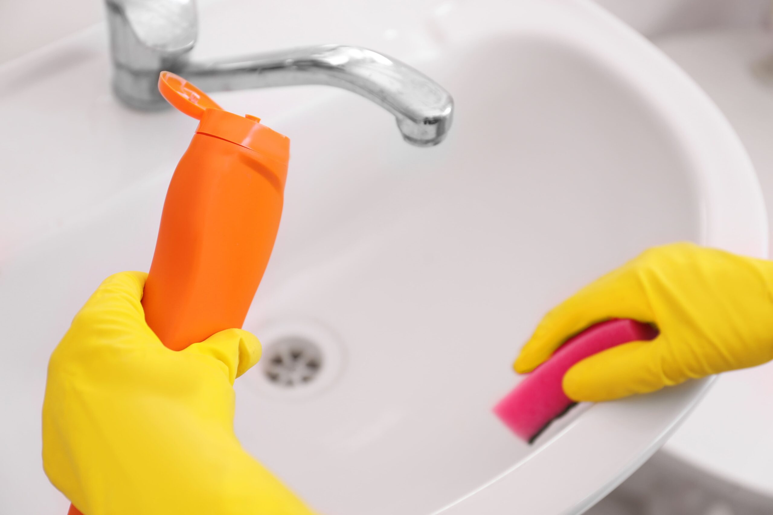 Person wearing yellow gloves cleaning a white sink with a sponge and an orange bottle of toilet bowl cleaner, safe to use for effective sink cleaning.