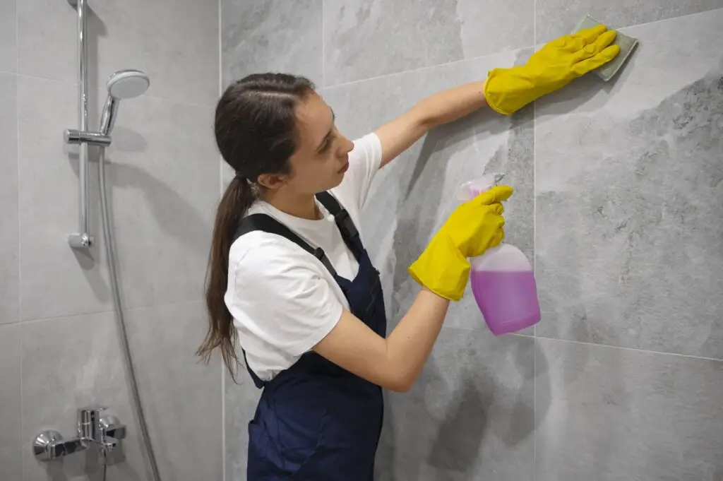 Woman wearing yellow gloves cleaning bathroom tiles with a spray bottle and sponge, tackling bathroom cleaning with ease.