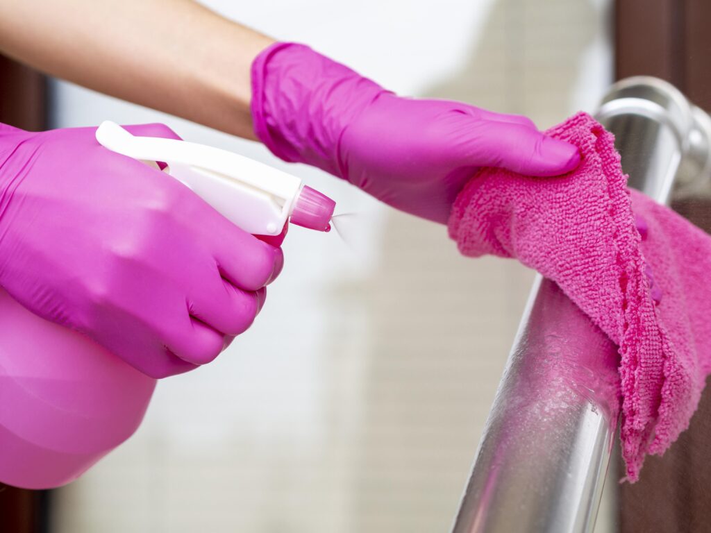 Hands in pink gloves deep cleaning a metal rail with a spray bottle and pink cloth.
