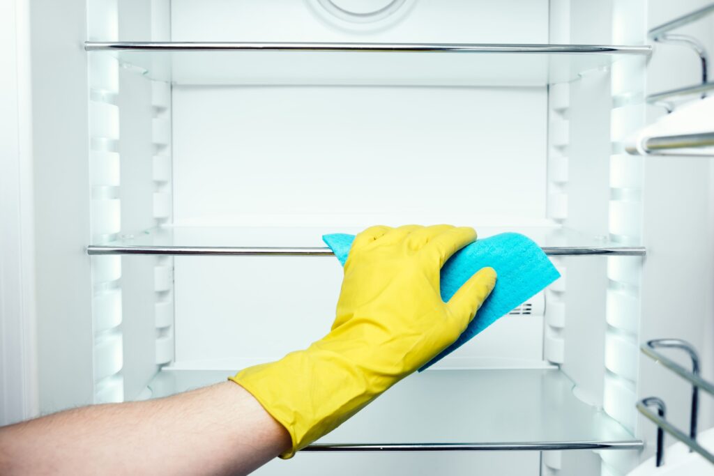 A hand in a yellow glove cleans an empty refrigerator shelf with a blue cloth, illustrating fridge cleaning as part of a step-by-step guide to clean inside of a fridge.