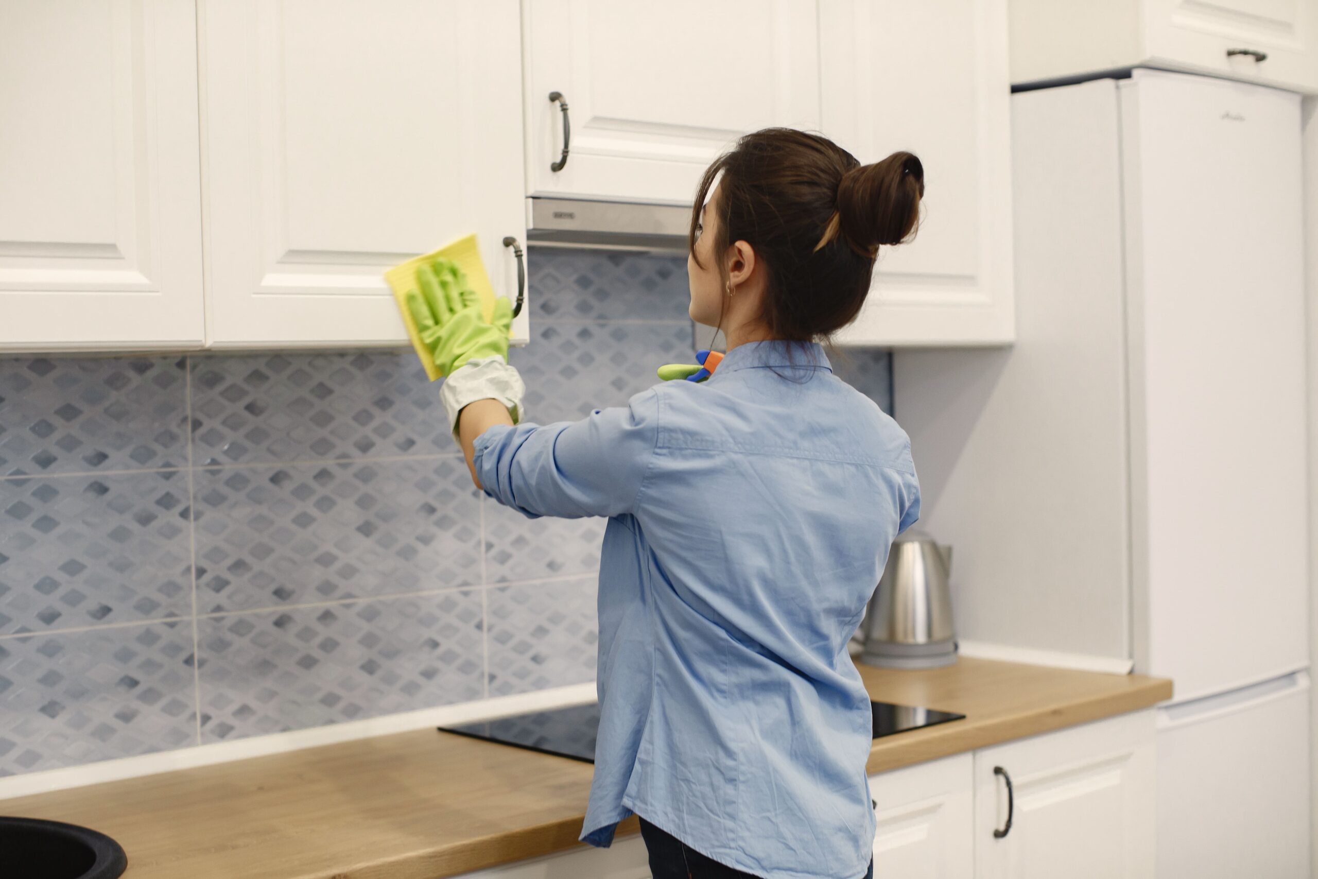 Woman wearing gloves cleaning kitchen cabinets with a sponge in a modern kitchen, showing how to clean inside cabinets and keep clean kitchen drawers spotless.