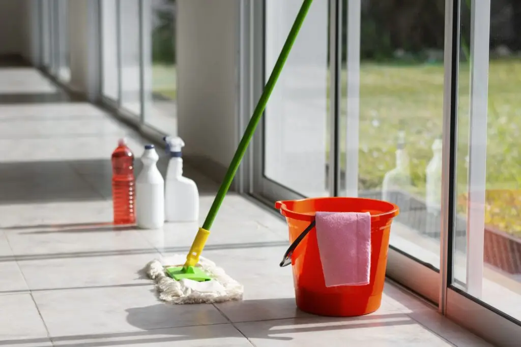 A mop, orange bucket, and cleaning supplies sit on a tiled floor by large glass windows in sunlight—everything you need for the best way to clean glass doors or tackle sliding door cleaning with ease.