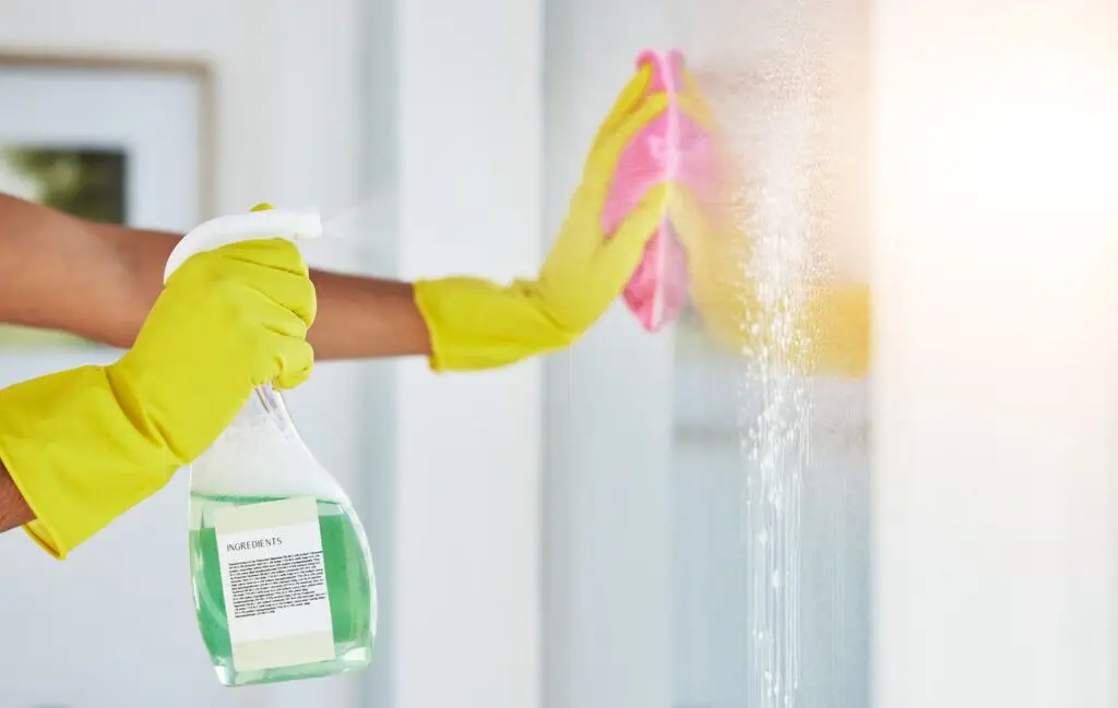 Person wearing yellow gloves sprays glass cleaner and wipes a window with a pink cloth, cleaning in sunlight for an effective cleaning comparison.
