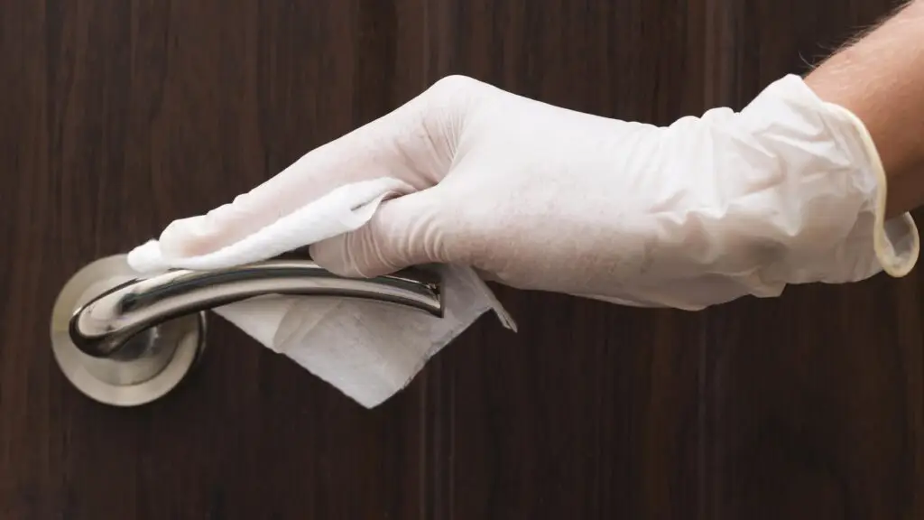 Gloved hand cleaning a door handle with a disinfectant wipe to sanitize home after illness.