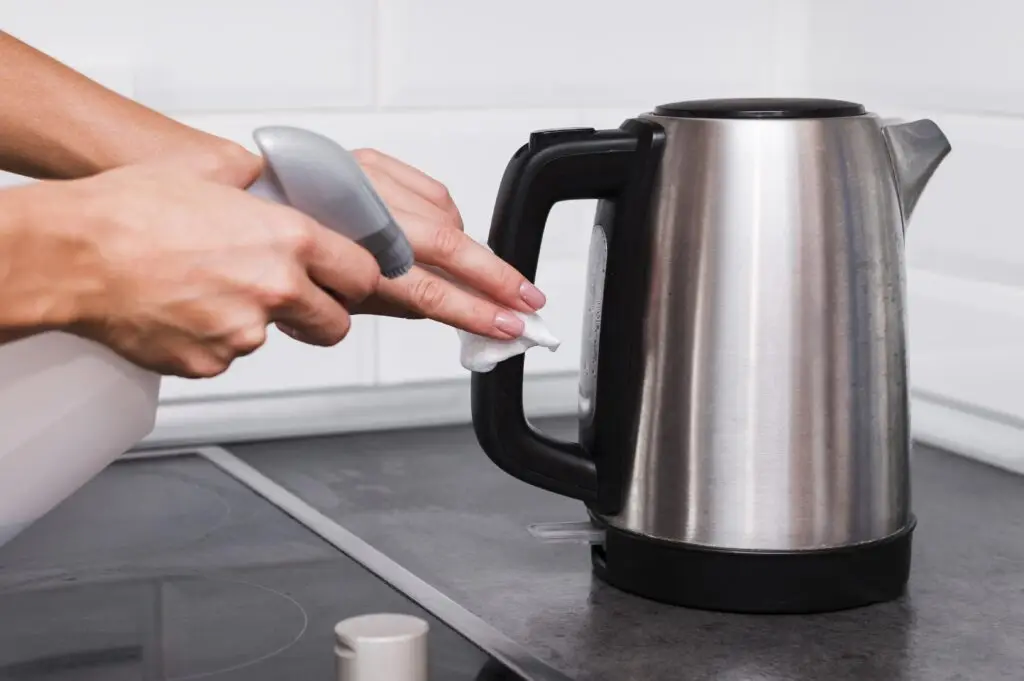 Hands cleaning a stainless steel electric kettle handle with a spray bottle and a cloth on a kitchen counter, demonstrating one of the best ways to clean small kitchen appliances for spotless, hygienic results.