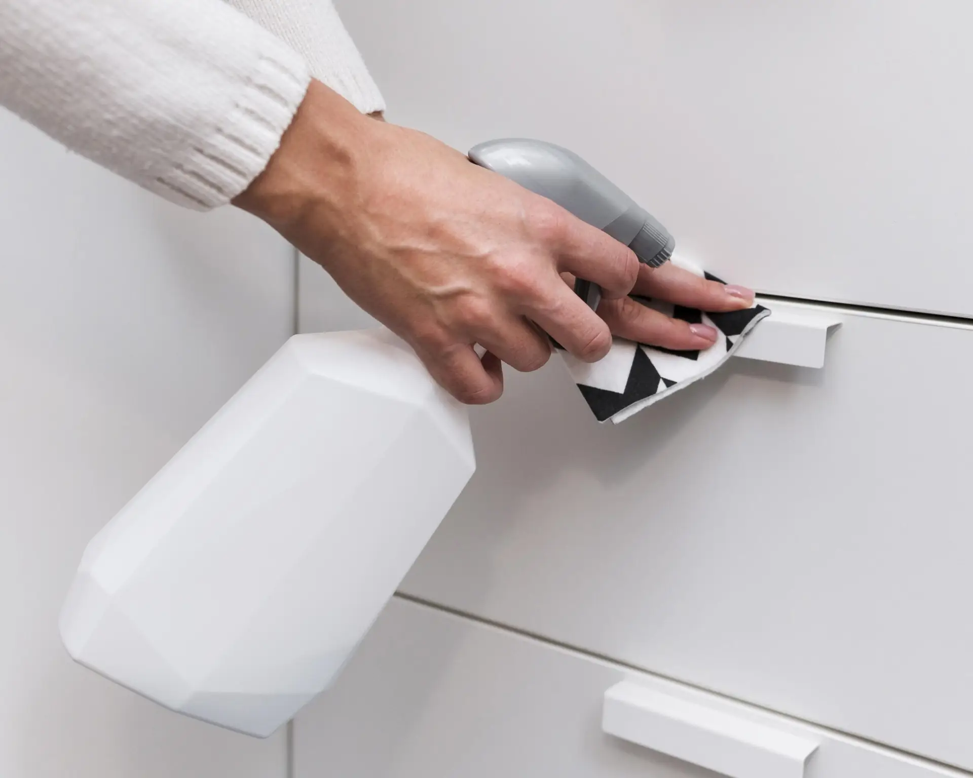 A hand sprays and wipes a white drawer handle with a cleaning cloth and bottle.
