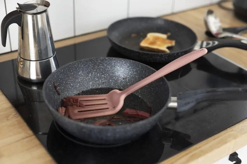 Nonstick cookware sits on the stove, featuring a pink spatula and food scraps, while toast crisps in another pan and a coffee pot brews nearby—just the essentials for any Pro Guide on how to remove burn marks and keep your kitchen running smoothly.