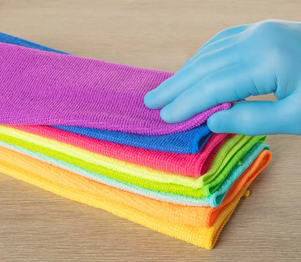 A gloved hand touches stacked, colorful reusable cleaning cloths on a wooden surface.
